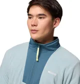 Buzo polar con cierre frontal completo de la marca Columbia. Presenta un diseño en color azul claro con paneles superpuestos en un tono azul más oscuro en la zona del pecho y cuello. Incluye bolsillos laterales con cierre, cuello alto y detalles en color amarillo neón en los tiradores de los cierres.