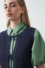 Chaleco tejido color verde musgo, con trama texturada, cuello a la base y cierre frontal con lazos. Presenta un ruedo con volado.