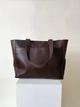 Cartera tipo tote de cuero graneado negro, con diseño clásico y textura uniforme. Cuenta con doble asa de hombro, bolsillo frontal con cierre magnético, bolsillo trasero con cierre metálico y compartimento principal con cierre. El interior está forrado y dispone de múltiples bolsillos organizadores.