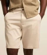 Bermuda masculina slim confeccionada en sarga de algodón con elastano, color beige, con bolsillos laterales tipo chino, bolsillos traseros embutidos con botón, trabillas en la cintura y cierre frontal con botón y cremallera.