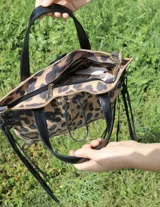 Cartera de cuero con estampado animal print de leopardo, con diseño de laterales acordonados con flecos largos en color negro. Cuenta con doble asa de mano y cierre superior con detalle de tiento.