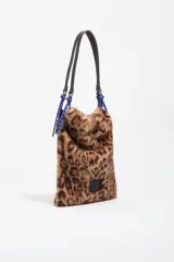 Bolso de hombro con estructura blanda y diseño ligero, confeccionado en tejido de pelo sintético con estampado de leopardo. Cuenta con asa de hombro de cuero y detalles de cordones trenzados en azul y rojo en las uniones. Incluye etiqueta de cuero con logo grabado en la parte frontal.
