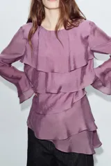 Blusa de cuello redondo y manga larga, color marrón oscuro. Presenta volantes en el frente y en los puños, y un bajo asimétrico. Cierre en la espalda con abertura y botón forrado.