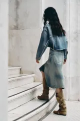 Falda midi de denim con lavado claro, tiro medio y cintura cruzada con cierre asimétrico de cremallera y botones metálicos. Presenta bolsillos delanteros y bajo asimétrico.