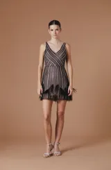 Vestido corto sin mangas en tono nude con capa de tul negro transparente superpuesta, adornado con líneas de pedrería brillante y detalles de plumas en el ruedo. Presenta escote en V pronunciado.