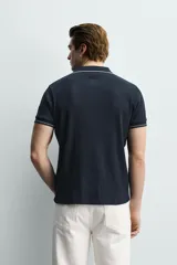 Remera polo de punto canalé color azul marino, de corte regular fit, con cuello solapa y cierre frontal de botones a presión. Cuenta con mangas cortas con ribetes blancos en los bordes del cuello y de las mangas.