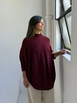 Poncho de punto color bordó con cuello alto y diseño de trenzas verticales en el frente. Presenta mangas tres cuartos con puños acanalados y un corte holgado con ruedo redondeado.
