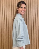 Blusa blanca de algodón con escote en V y cuello mao. Presenta mangas largas abullonadas con puños acampanados y fruncidos.