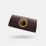 Bolso rígido alargado de tela color marrón, con solapa y aplique central de piedra de resina sobre base metálica dorada. Cuenta con asa de cadena desmontable.