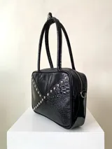 Cartera rígida negra con detalles en rojo y tachas plateadas. Tiene herrajes y cierre metálico plateado marca YKK, asa fina para llevar al hombro o en la mano y correa larga extra regulable. El interior es forrado, tiene un bolsillo con cierre enmarcado en cuero, y un segundo bolsillo plaqué.