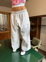 Pantalón de jogging gris de corte baggy, confeccionado en algodón. Presenta cintura elástica con cordón largo ajustable, botones en la parte inferior para ajustar la botamanga y un diseño artístico que combina estampa en plastisol con bordado en relieve sobre el muslo izquierdo.
