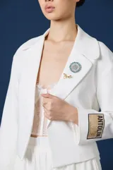 Saco corto de color blanco con solapas clásicas, mangas largas y detalle de parche rectangular con texto en el puño izquierdo. Presenta una silueta estructurada y elegante.