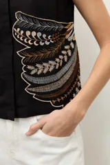 Blusa negra sin mangas, con cuello redondo y cierre frontal de botones ocultos. Presenta un aplique bordado con diseño abstracto en el lateral inferior.