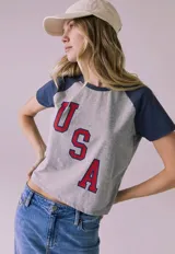 Remera gris de manga corta con mangas ranglán azul marino y estampa frontal con las letras "USA" en rojo con borde azul.