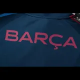 Camiseta de fútbol azul marino con mangas raglán, cierre de cremallera corto en el cuello, escudo del FC Barcelona bordado en el pecho y detalles en azul y rojo en mangas y laterales.