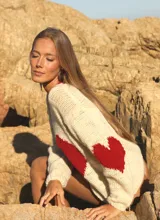 Sweater tejido de punto color marfil con cuello redondo y diseño frontal de corazón rojo. Cuenta con detalles en rojo en el borde del cuello y las mangas.
