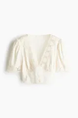 Cropped top de color beige claro, con escote en V pronunciado, mangas cortas abullonadas y cierre frontal con pequeños botones. Presenta detalles de encaje floral en el escote y los puños.