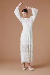 Vestido largo color crudo de encaje, con mangas largas abullonadas, cuello redondo y falda con paneles traslúcidos.