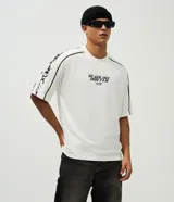 Remera de corte boxy y silueta amplia, color blanco con cuello redondo. Presenta estampa frontal con texto y gráficos tribales en las mangas.