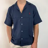 Camisa de hombre azul marino de muselina con manga corta y corte oversize. Presenta cuello tipo cubano, cierre frontal con botones blancos y un pequeño logo bordado en rojo en el pecho.
