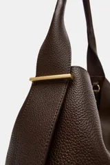 Bolso tipo saco de gran tamaño en cuero marrón oscuro, con textura granulada. Presenta un asa de hombro ancha y detalles metálicos dorados en la unión del asa. Incluye un bolsillo interior extraíble y cierre mediante botón imantado.