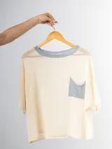 Remera tejida en hilo italiano color crema, con cuello redondo y bolsillo frontal gris. De caída relajada y silueta amplia.