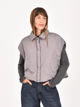 Chaleco puffer corto de color gris, con cuello camisero y cierre frontal oculto. Presenta un diseño holgado y sin mangas.