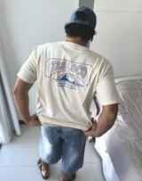 Remera de manga corta color beige jaspeado, con un estampado gráfico en la espalda que incluye el texto "TAS LOCO" y "SUNNY WITH A CHANCE OF STOKE" con un diseño de una persona en el agua con una ballena.