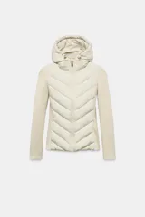 Cazadora de mujer con diseño híbrido, presenta un panel frontal acolchado con costuras en chevron y mangas y laterales en tejido liso. Cuenta con cuello alto, capucha ajustable con cordones elásticos y cierre frontal mediante cremallera.