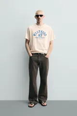 Camiseta relaxed fit de algodón color beige claro, con cuello redondo y mangas raglán cortas. Cuenta con estampado frontal en tono azul con efecto desgastado que incluye texto y el número 87.