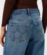 Pantalón de jean estilo wide leg con cintura media, cierre frontal con botón y bolsillos funcionales. Presenta un diseño de corte amplio y recto, con bordado de estrellas en los bolsillos traseros.