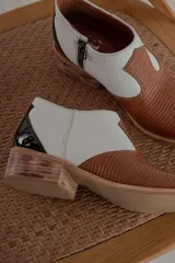Botinetas de cuero con diseño bicolor, combinando cuero texturizado marrón en la puntera y cuero blanco granulado en los laterales. Presenta un detalle de charol negro en el talón y taco bajo de madera.