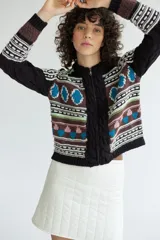 Campera de punto jacquard con lana merino, diseño con cierre frontal, cuello redondo y detalles de trenzas en los laterales. Presenta un patrón geométrico multicolor con tonos marrones, negros, azules y verdes.