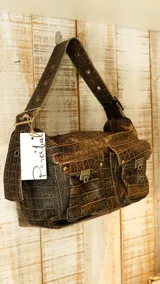 Bolso de hombro color beige con textura de cuero grabado tipo cocodrilo. Presenta dos bolsillos frontales con solapa y cierres metálicos, y una correa ancha ajustable con ojales metálicos.