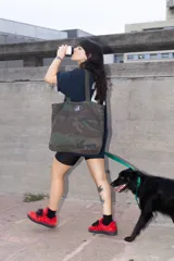 Bolso tote de tela con estampado de camuflaje en tonos verdes y marrones. Tiene asas de hombro y un logo ovalado con letras blancas en el centro.
