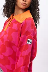 Buzo de tejido de punto con diseño geométrico en tonos fucsia y rojo, cuello tipo polo con cierre de botones y puños en color naranja contrastante. Presenta un corte holgado y oversize.