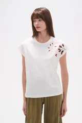 Remera blanca de algodón con mangas cortas y cuello redondo. Presenta un bordado floral en tonos rosados y rojizos en la zona del hombro derecho.