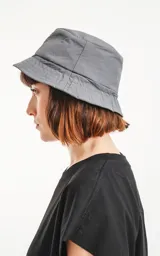Gorro tipo bucket de color gris oscuro (carbón), con ala ancha y un pequeño parche negro con un logo bordado en blanco en el frente.