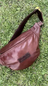 Riñonera de cuero vacuno color camel, con cierre principal y tirador de cuero, correa regulable y parche con logo grabado en el frente.