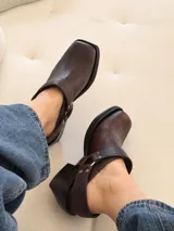 Zueco de cuero marrón con textura envejecida, diseño de punta cuadrada y taco bajo. Presenta una tira trasera con hebilla metálica y detalles de tachas plateadas.