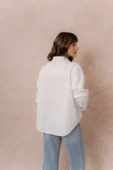 Camisa de denim azul con bordados florales en tonos amarillos y celestes sobre los hombros y el pecho. Presenta cuello clásico, cierre frontal con botones y mangas largas con puños.