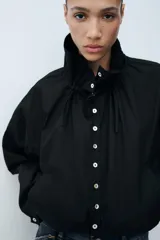 Cazadora de algodón negra con cuello subido ajustable con lazadas, manga larga, bolsillos de vivo en los laterales y bajo ajustable con cordones. Cierre frontal con botones.