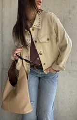 Sobrecamisa beige de gabardina con tachas metálicas decorativas en el cuello y la solapa frontal. Cuenta con bolsillos con solapa en el pecho y cierre frontal con botones.