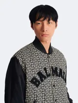 Campera estilo varsity de Balmain con cuerpo estampado con monograma geométrico y mangas negras lisas. Presenta cierre frontal con botones a presión, cuello, puños y ruedo acanalados, y el nombre de la marca aplicado en letras de felpa en el pecho.