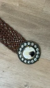 Cinto de cuero trenzado color marrón chocolate con hebilla circular grande de metal plateado, decorada con tachas esféricas y un centro de pelo natural.