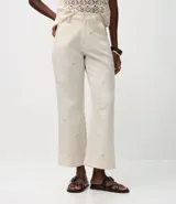 Pantalón de mujer color blanco roto, de corte recto y tiro alto, con un largo cropped y ligero ensanchamiento en el bajo. Presenta detalles bordados florales sutiles en la tela y bolsillos.