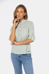 Camisa blanca de manga larga con cuello y tapeta de medio botón. Presenta dos bolsillos frontales con tapa y botones metálicos. Las mangas tienen presillas para sujetar el puño enrollado.
