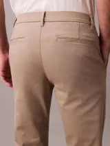 Pantalón chino de corte slim, confeccionado en tejido elástico de algodón orgánico. Presenta cierre de botón y cremallera, bolsillos laterales y bolsillos traseros ribeteados.