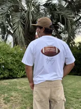 Remera blanca de algodón con estampado de un balón de fútbol americano y la frase "Together, we are America" en rojo. Incluye una firma en negro debajo del estampado.