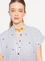 Túnica blanca con rayas verticales azules y estampado de hojas negras, cuello mao, mangas cortas amplias y cierre frontal con botones.